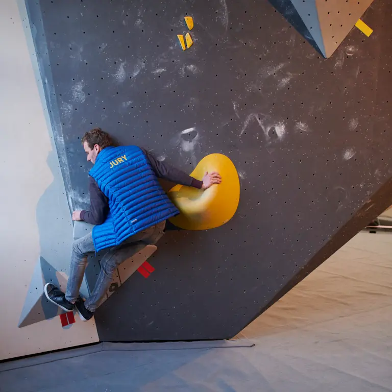 Iemand die een blauw vest draagt met het opschrift ‘JURY’ op de rug, is binnen aan het boulderen en houdt zich vast aan een grote gele klimgreep op een overhangende wand. De omgeving lijkt een klimhal of een wedstrijd te zijn.